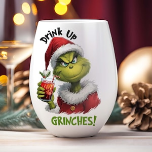 Może przedstawiać: Biały kieliszek do wina bez nóżki z motywem Grincha. Grinch ma na sobie czapkę Świętego Mikołaja i czerwony płaszcz, trzymając koktajl. Tekst na szkle brzmi "Drink Up Grinches!" Świąteczny motyw.