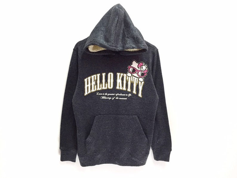 May include: A gray hooded sweatshirt with a Hello Kitty graphic on the front. The graphic features Hello Kitty wearing sunglasses and the text "Hello Kitty" in gold lettering. The text "Love is the greatest refreshment in life. The blessing of the moment." is printed below the graphic.