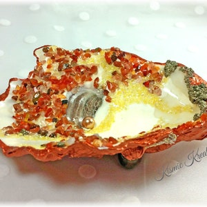May include: A white seashell with a copper-coloured rim filled with small red and yellow stones, a small white shell, and a pearl.