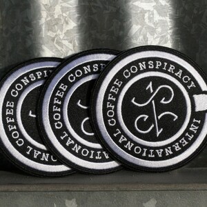 May include: Three black and white embroidered patches with the text "Coffee Conspiracy International" and a stylized coffee cup design.