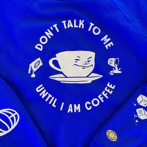 Op de afbeelding: Blauw sweatshirt met witte tekst die "Praat me niet aan tot ik koffie heb" zegt met een cartoon koffiekopje en een gemorst kopje koffie.