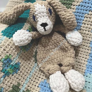 May include: A handmade, crocheted dog plushie in shades of tan, white, and black. The dog has floppy ears, a white snout, and white paws. The plushie is lying on a crocheted blanket with blue, green, and tan geometric patterns. The text "Custom Made by Hooked on the Sea" is visible.