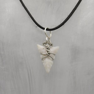 Puede incluir: Un colgante de punta de flecha blanca envuelto en alambre plateado, colgando de un collar de cordón negro.