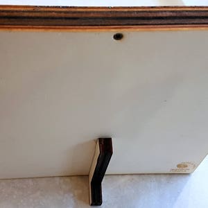 Puede incluir: La parte posterior de un marco de fotos de madera blanca con un soporte de madera marr&oacute;n oscuro. El marco tiene un peque&ntilde;o tornillo en la esquina superior derecha.