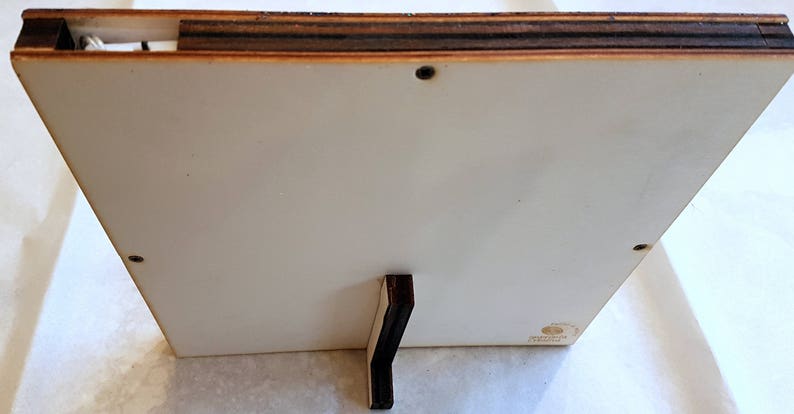 Puede incluir: La parte posterior de un marco de fotos de madera blanca con un soporte de madera marr&oacute;n oscuro. El marco tiene un peque&ntilde;o tornillo en la esquina superior derecha.