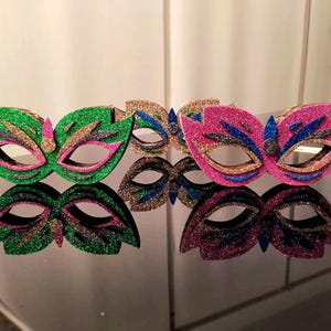 May include: Glittering masquerade masks in green, gold, and pink. The masks have a leaf-like design with cut-out eye holes and are decorated with glitter and colourful accents. The masks are displayed on a reflective surface.