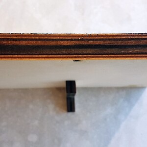 Puede incluir: Primer plano de la parte inferior de un marco de madera. El marco est&aacute; hecho de madera contrachapada con una chapa de madera oscura. Una peque&ntilde;a pieza de metal es visible en la parte superior central del marco.