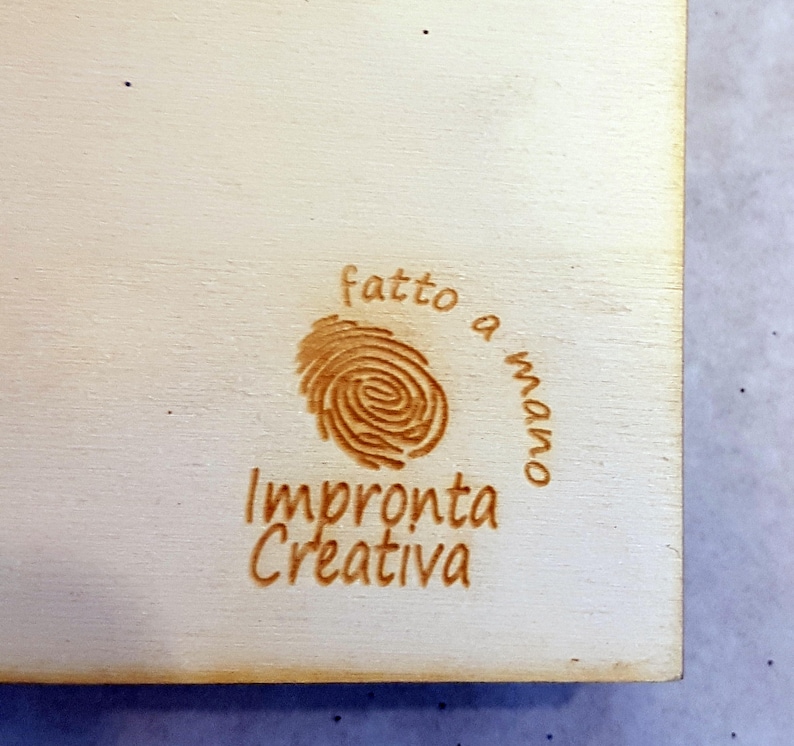 Puede incluir: Un letrero de madera con un dise&ntilde;o de huella digital marr&oacute;n y el texto "fatto a mano" (italiano para "hecho a mano") e "Impronta Creativa" (italiano para "Huella Creativa").