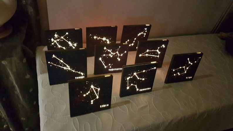 Puede incluir: Doce placas de madera con constelaciones y sus nombres, iluminadas con luces blancas. Las placas est&aacute;n dispuestas en fila sobre una superficie blanca.