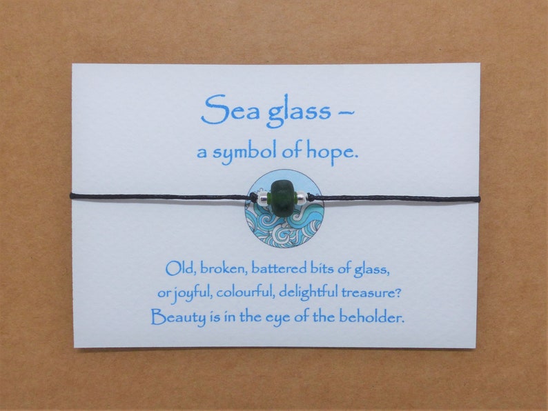 May include: A black string bracelet with a green sea glass bead and a silver bead. The bracelet is on a white card with the text "Sea glass - a symbol of hope. Old, broken, battered bits of glass, or joyful, colourful, delightful treasure? Beauty is in the eye of the beholder."