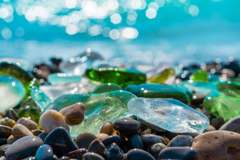 May include: Close-up of smooth, colorful sea glass and gray pebbles on a beach. The sea glass is various shades of blue, green, and clear. The pebbles are smooth and gray.
