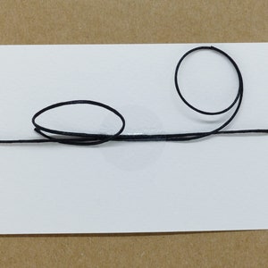 May include: A white card with a black string tied around it in a simple design. The string is tied in a knot on the top of the card and then forms two loops, one on each side of the card.