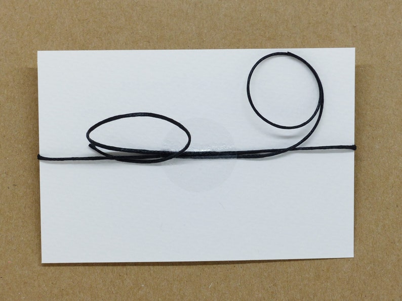 May include: A white card with a black string tied around it in a simple design. The string is tied in a knot on the top of the card and then forms two loops, one on each side of the card.