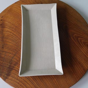 Può includere: Un vassoio da portata rettangolare in ceramica grigio chiaro con lati leggermente curvi e una finitura opaca liscia. Il vassoio presenta una base piatta e bordi rialzati, offrendo un'estetica pulita e neutra per servire vari alimenti.
