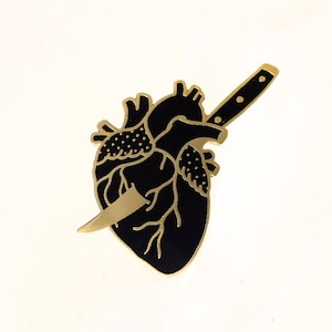 May include: A black and gold enamel pin depicting a heart pierced by a knife. The heart is detailed with veins and the knife has a gold handle.