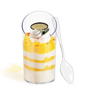 Op de afbeelding: Transparante plastic dessertbekers met een geel en wit gelaagd dessert, bedekt met slagroom en rode sprinkles. De bekers hebben een lichte bocht aan de bovenkant en worden geleverd met een transparante plastic lepel.