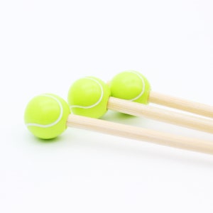 May include: Three novelty tennis ball-shaped items with light green spheres and white stripes, attached to wooden dowels. The items are arranged diagonally against a white background, suggesting a playful or sports-themed product.