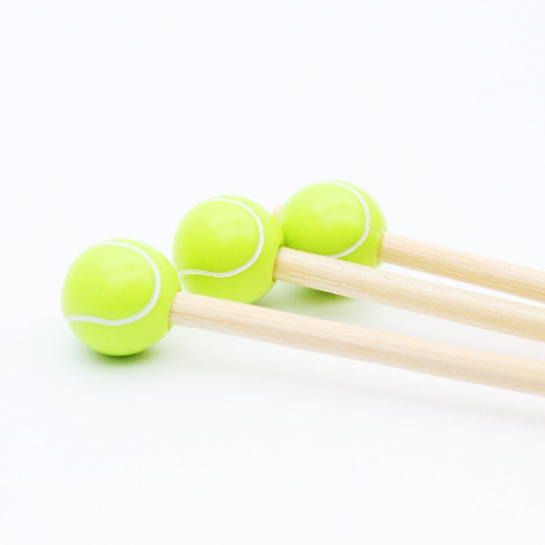 May include: Three novelty tennis ball-shaped items with light green spheres and white stripes, attached to wooden dowels. The items are arranged diagonally against a white background, suggesting a playful or sports-themed product.