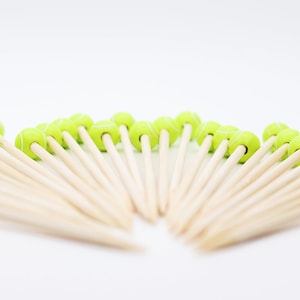May include: A collection of wooden cocktail picks, each topped with a small, green tennis ball design. The tennis balls have white stripes, and the picks are arranged in a fan shape against a white background. These are ideal for a tennis-themed party.
