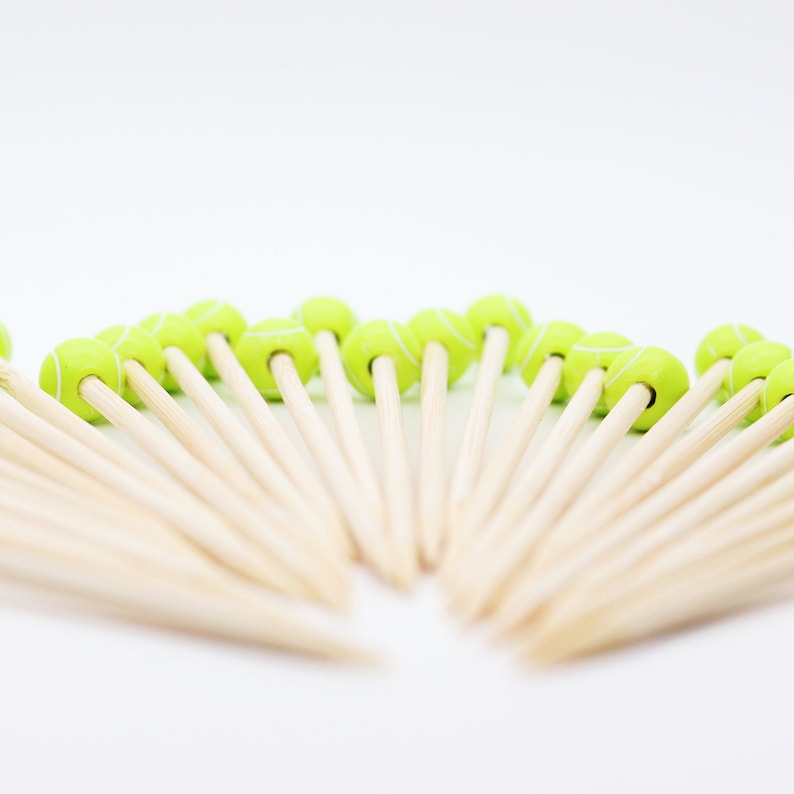 May include: A collection of wooden cocktail picks, each topped with a small, green tennis ball design. The tennis balls have white stripes, and the picks are arranged in a fan shape against a white background. These are ideal for a tennis-themed party.