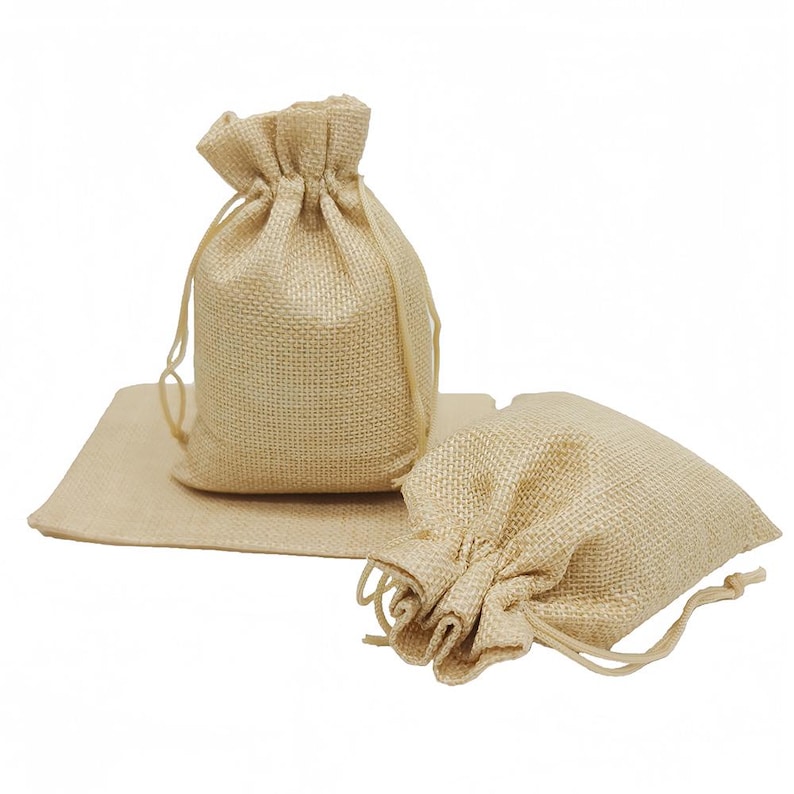 May include: Three beige burlap drawstring bags are shown. One bag stands upright, another lies on its side, and the third rests on a matching burlap mat. The bags have a textured weave and a natural, neutral colour.