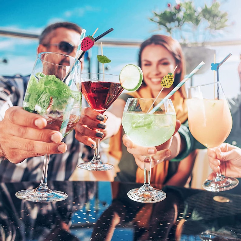 May include: A group of people holding up cocktail glasses filled with colorful drinks. The drinks are garnished with fruit and decorative skewers. The glasses are clear, and the drinks are various shades of red, green, and yellow.