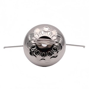 May include: A shiny, silver-colored tea infuser with a spherical body and a decorative floral-like pattern around the top. It has a small handle and two thin, straight arms extending from the sides.