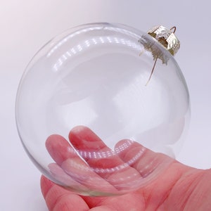 Könnte beinhalten: Ein transparenter Weihnachtsbaumschmuck aus Glas mit einem silbernen Metallverschluss. Der Schmuck wird in einer Hand gehalten.
