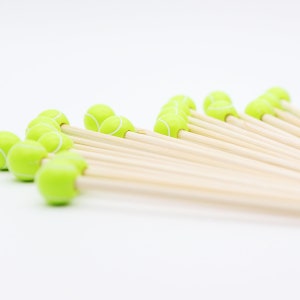 May include: A collection of wooden skewers topped with small, bright green tennis ball decorations. The tennis balls have white stripes and are arranged in a diagonal pattern against a white background. These skewers are ideal for appetizers or party snacks.