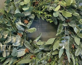 May include: A fresh, green Christmas wreath composed of various evergreen branches and eucalyptus leaves. The wreath is adorned with star anise and other natural elements, creating a rustic and festive holiday decoration. The wreath is perfect for adding a touch of nature to any home.