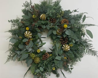 May include: A circular Christmas wreath composed of various green foliage, including pine and eucalyptus. The wreath is adorned with pine cones, dried orange slices, and yellow spherical flowers. The wreath is displayed against a plain white background.