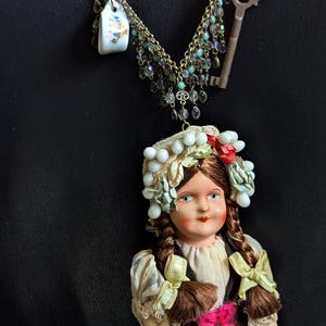 May include: A vintage porcelain doll with brown hair in braids, wearing a white dress with a brown vest and a red skirt. The doll is wearing a white and green floral crown and is adorned with a necklace featuring a key, a small teacup, and beads.