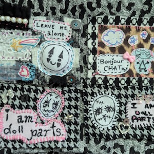 May include: A textile collage featuring four distinct fabric patches on a black and white patterned background. The patches include hand-stitched embroidery, white fabric with black ink drawings, pink felt text reading "I am doll parts," and "Leave me alone," plus small decorative buttons, beads, and a pink bow.