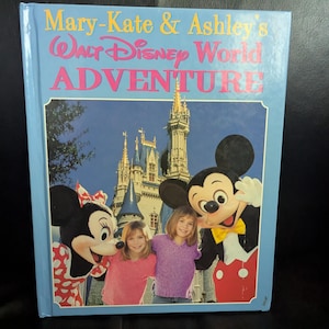 May include: A hardback book titled "Mary-Kate & Ashley's Walt Disney World Adventure" with a light blue cover. The cover features Minnie Mouse, Mickey Mouse, two girls, and Cinderella's castle. The title is in pink and yellow.