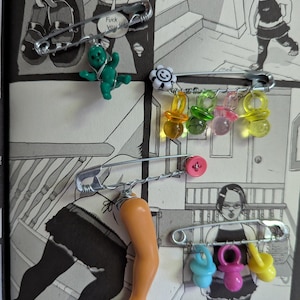 May include: A collection of safety pins adorned with various small charms and objects. The charms include a green plastic baby, a white and black flower, several colorful pacifiers, a pink button, and a doll arm. The background is a black and white comic strip.