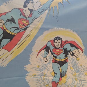 May include: Light blue fabric featuring a repeating pattern of Superman in action poses. The illustrations show Superman in his classic costume with a red cape, blue suit, and the iconic "S" shield. Yellow and white accents create a dynamic background.