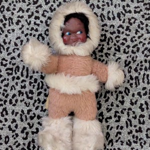 Può includere: Una bambola vintage vestita con un completo in finta pelliccia rosa e bianca, tra cui un parka con cappuccio, guanti e stivali. La bambola ha una carnagione scura ed è ambientata su uno sfondo con stampa leopardata grigia e nera.