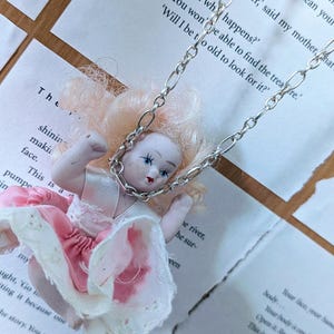 May include: A vintage doll pendant necklace. The doll has a porcelain face, pink hair, and a pink and white dress. The necklace chain is silver-coloured. The background is a book with printed text.
