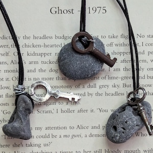 May include: Three necklaces with grey stone pendants and antique silver keys. The necklaces are made with black cord and are hanging on a book with the text "Ghost - 1975".