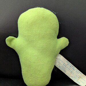 May include: A lime green plush toy in the shape of a cactus with two small arms. A white tag is attached to the toy. The tag has text printed on it. The toy is made of a soft, fuzzy material.