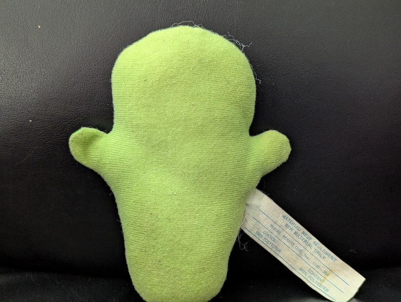 May include: A lime green plush toy in the shape of a cactus with two small arms. A white tag is attached to the toy. The tag has text printed on it. The toy is made of a soft, fuzzy material.