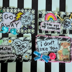 May include: Four rectangular fabric patches featuring black and white houndstooth patterns. The patches are decorated with embroidered text including "GO AWAY," "little ray of sunshine," "MAYBE tomorrow," and "i don't like You," alongside colorful appliques.