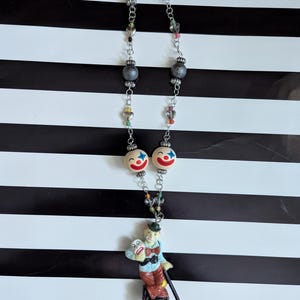 May include: A silver chain necklace with grey, clear, and colourful beads. The necklace features two clown face beads and a pendant of a clown holding a mask.