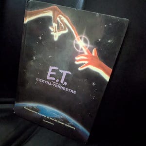 May include: A hardcover book titled "E.T. L'EXTRA-TERRESTRE" with a space-themed cover. The cover art features two hands reaching towards each other with a glowing light in between. The book is a collection of photographs from the Steven Spielberg film.