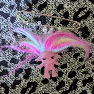 Puede incluir: Una figura rosa con pelo colorido está unida a un imperdible grande. La figura tiene ojos negros y está de pie. El pelo es una mezcla de rosa, amarillo y azul. Pequeñas cuentas blancas también están unidas al imperdible.