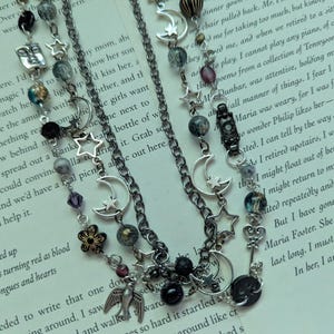 May include: A multi-layered necklace with moon and star charms, decorative beads, and pendants. The dark silver chain necklace is displayed on a book page.
