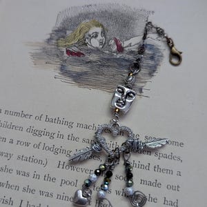 May include: A silver-toned charm with a face pendant, feathers, hearts, and beads. The charm is attached to a chain with a clasp. The background is a vintage book page with an illustration of a girl in water.