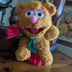 May include: A plush brown teddy bear wearing a green and red scarf. The bear has a pink nose and a wide, open mouth.