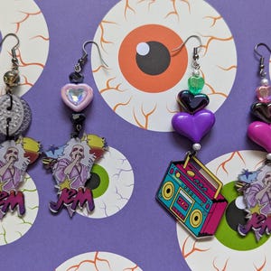 Peut inclure: Une collection de boucles d'oreilles pendantes colorées. Une paire présente un cœur rose, un cœur noir et un cœur violet. Une autre paire a un cœur rose avec une gemme et un disque transparent. Chaque boucle d'oreille a un motif JEM.