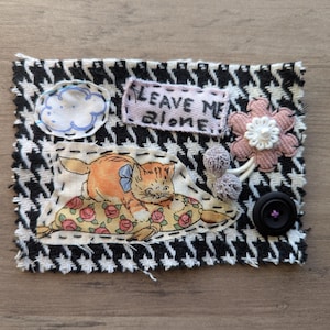May include: A fabric patch with a black and white houndstooth pattern. It features an orange cat on a floral pillow, a cloud, a pink flower, two gray pom-poms, a black button, and a banner that reads "LEAVE ME ALONE."